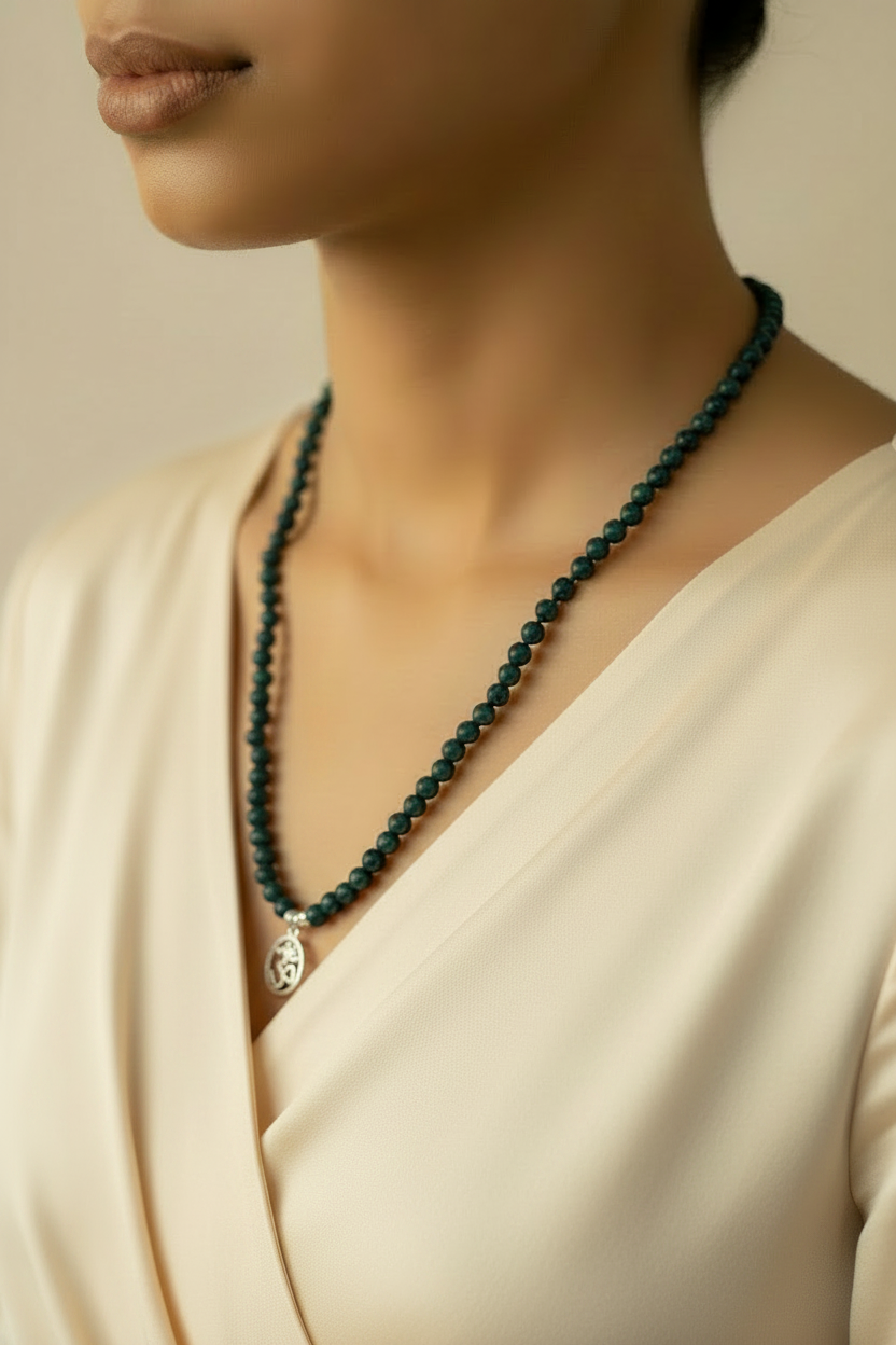 Person wearing a dark green beaded necklace with a silver pendant on a neutral background